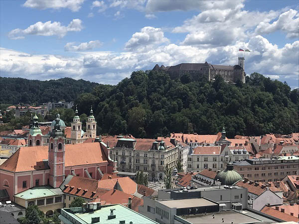 Een kijk op het kasteel van Ljubljana op de achtergrond en op de voorgrand de rode Franciscuskerk van de Maria Boodschap met daarachter de 2 uitstekende torens van de Sint-Nicolaaskathedraal<